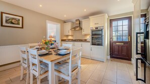 A modern kitchen with white cabinetry, a dining area with a wooden table set for four, and a large window providing natural light. - 3 Bed in St. Dogmaels (oc-y30253) (Saint Dogmaels)