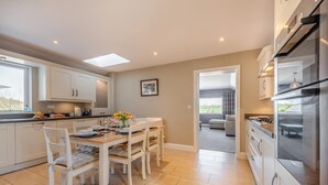 A modern kitchen with white cabinets, a dining area with a wooden table and chairs, and a view into a living room. - 3 Bed in St. Dogmaels (oc-y30253) (Saint Dogmaels)