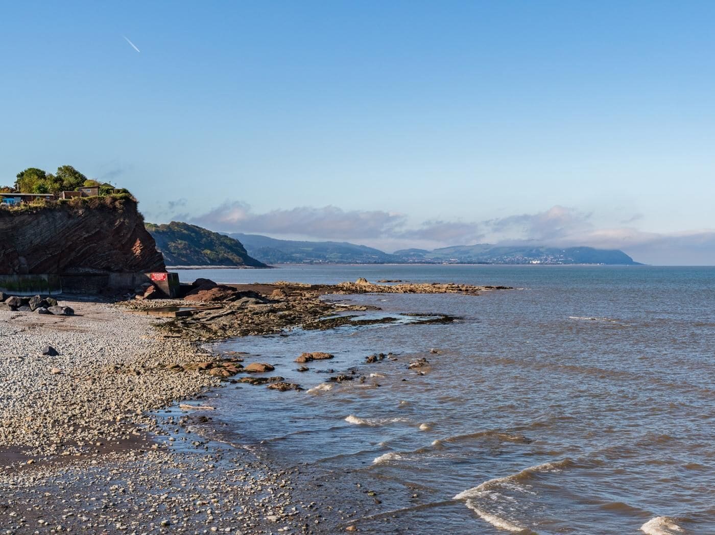 A rocky beach with a cliff, calm sea, and distant mountains.