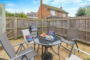 A patio with a round table, four chairs, a grill, and a wooden fence. - 3 Bed in Watchet (oc-c29959) (Watchet)