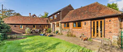 A two-story house with a brick exterior and a tiled roof. There is a small garden with a lawn and a wooden table.