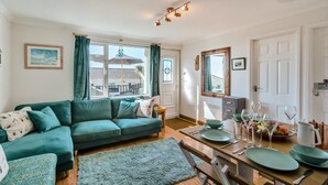 A living room with a green sofa, a dining table set with plates and glasses, a small side table, and a window with curtains. - 2 Bed in Mullion (oc-k7968) (Mullion)