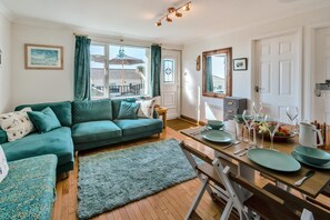 A living room with a green sofa, a dining table set with plates and glasses, a small side table, and a window with curtains. - 2 Bed in Mullion (oc-k7968) (Mullion)