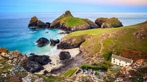 A coastal landscape with rocky outcrops, a sandy beach, and a white building with a garden. - 2 Bed in Mullion (oc-k7968) (Mullion)