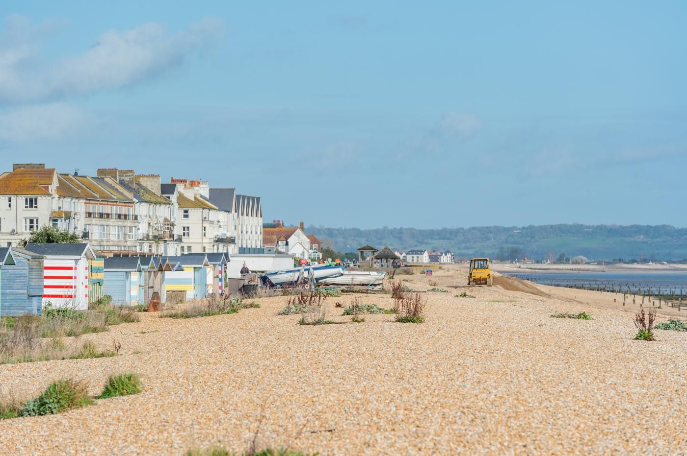 A pebbled beach with beach huts, a parked yellow vehicle, and houses in the background.