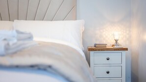 En pænt redt seng med hvide sengetæpper, et natbord med en lampe og en lille skuffe.