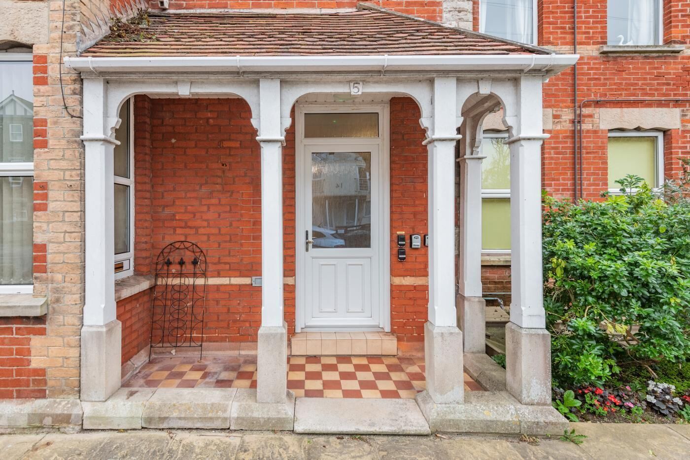 Une maison en briques rouges avec un porche blanc, un sol à carreaux et le chiffre « 5 » au-dessus de la porte.