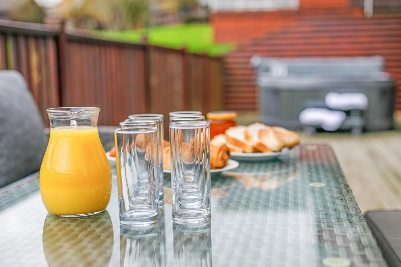 A glass table with a pitcher of orange juice and several glasses, a plate with croissants, and a grill in the background.