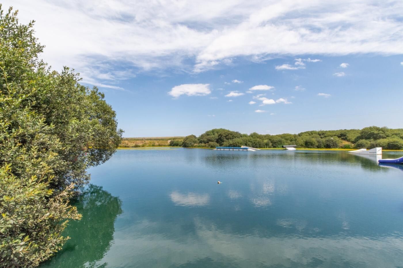 Ein ruhiger See mit Bootssteg, umgeben von dichtem Grün und Bäumen unter blauem Himmel.