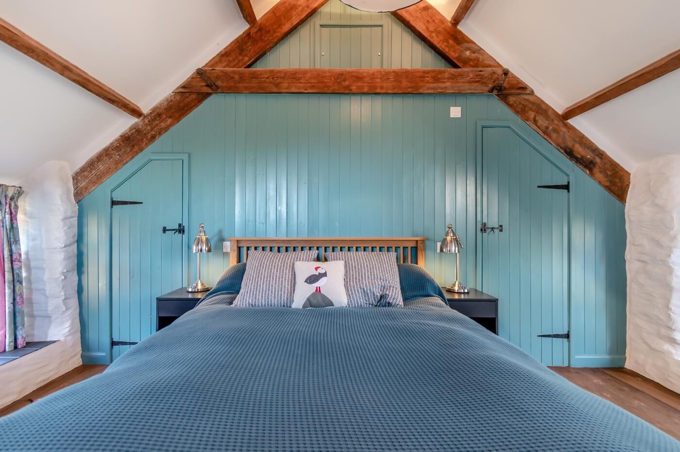 A bedroom with a sloped ceiling, a bed with a wooden headboard, two bedside tables with lamps, and a window with curtains.
