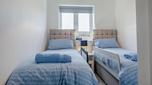 A bedroom with two beds, each with a headboard and a window. There is a small table between the beds. - 3 Bed in Totland Bay (oc-s30266) (Totland)