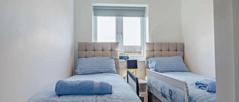 A bedroom with two beds, each with a headboard and a window. There is a small table between the beds.