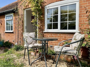 Een bakstenen huis met een groot raam, een terras met een ronde tafel en twee stoelen, en een tuin met planten en bloemen.