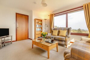 A living room with a sofa, a coffee table, a bookshelf, a television, and a large window with curtains. - 2 Bed in Ardvasar (oc-l30278) (Ardvasar)