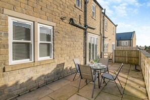 Une maison en briques dotée d’un patio comprenant une table et des chaises, une clôture en bois et une fenêtre avec des stores.
