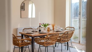 Uma sala de jantar com uma mesa posta para quatro pessoas, rodeada por cadeiras de vime, um espelho na parede e uma janela com vista.