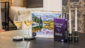 Ein gemütliches Zimmer mit einem Bett, einem Tisch mit zwei Weingläsern, einer Kerze und einem Buch mit dem Titel „Let's Get Deep Questions For Couples“.