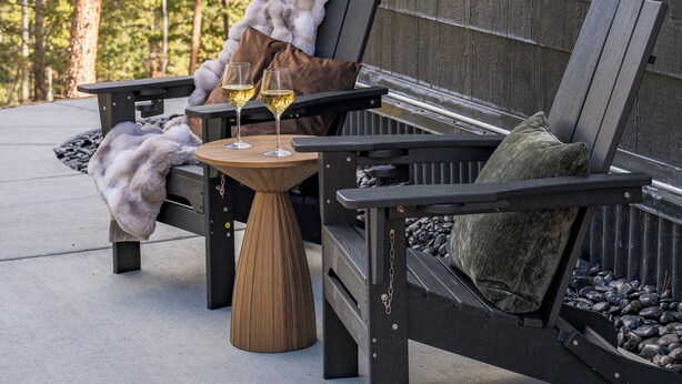 Ein gemütlicher Außenbereich mit einer hölzernen Bank und einem kleinen Rundtisch, umgeben von einer Holzterrasse und einem Wald im Hintergrund.