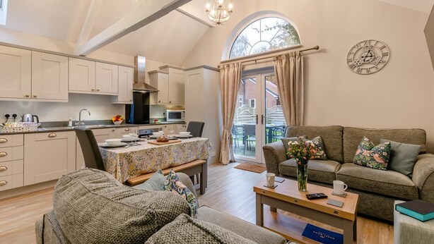 A cozy living room with a sofa, a coffee table, and a dining area with a table set for two. A kitchen with modern appliances and cabinets is visible in the background. - 3 Bed in Wilberfoss (oc-t30301) (Wilberfoss)