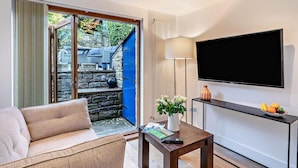 Ein Wohnzimmer mit einer Couch, einem Couchtisch mit Vase und Fernbedienung, einem Fernseher an der Wand und Blick auf einen Garten durch eine Glastür.