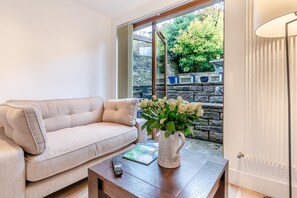 A living room with a beige sofa, a coffee table with a vase of flowers, a remote control, and a view of a stone wall and greenery outside. - 2 Bed in Hayfield (oc-t30237) (Hayfield)