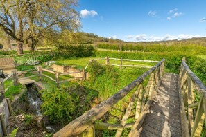 A wooden bridge over a stream, surrounded by lush greenery and a stone building.