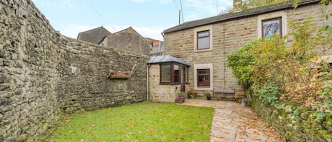 A stone wall, a grassy yard, and a stone pathway leading to a building with a window and a door.