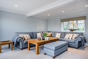 A modern living room with a grey sectional sofa, a wooden coffee table, and a matching ottoman. There is a small side table and a window with a sailboat decoration.