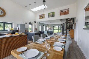 Eine moderne Küche mit Essbereich, gedeckt für eine Mahlzeit, ausgestattet mit einem langen Holztisch, hängenden Pendelleuchten und einer Treppe, die zu einer oberen Ebene führt.