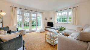 Amplia sala de estar con sofá, sillones, una mesa de centro y televisión. Amplias ventanas con cortinas permiten luz natural y vistas al exterior.