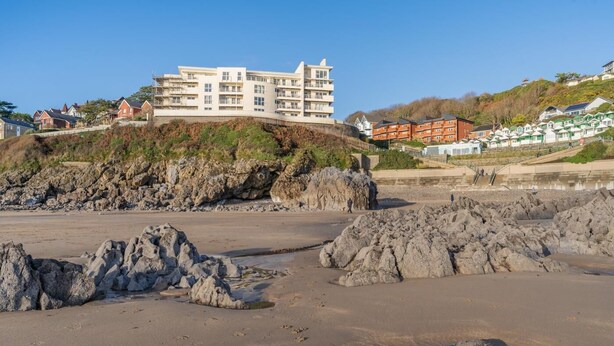 Um hotel à beira-mar com vários andares, uma praia de pedras em primeiro plano e um morro com edifícios residenciais.