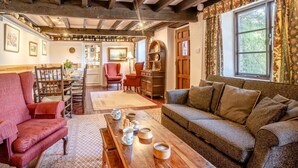 A cozy living room with exposed wooden beams, a dining area with wooden furniture, and a view through a window with floral curtains.
