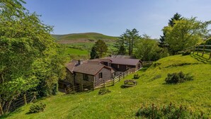 A rustic cabin with a wooden exterior, surrounded by lush greenery and a hilly landscape.