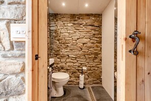 A small, stone-walled bathroom with a toilet, a hand dryer, and a wooden door with a metal handle.