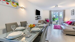 A modern dining area with a glass table, chairs, and a fruit bowl. There is a television mounted on the wall and a sofa with cushions. - 3 Bed in St. Ives (oc-c30315) (Carbis Bay)