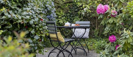 Un patio jardín con mesa y dos sillas, rodeado de follaje exuberante y plantas en flor.