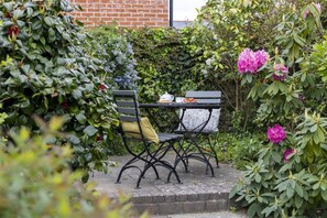 A garden patio with a table and two chairs, surrounded by lush greenery and flowering plants. - 1 Bed in Bridport (oc-p30346) (Bridport)