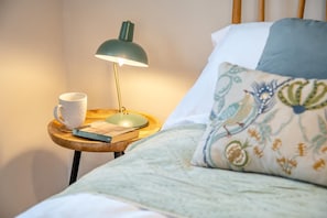 Ein Bett mit einem floral gemusterten Kopfkissen, ein Nachttisch mit Lampe, Tasse und Buch.