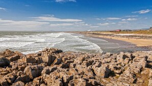 A rocky coastline with waves crashing onto the shore, a sandy beach, and a distant building. - 3 Bed in Pontardawe (oc-c30415) (Cilybebyll Pontardawe)