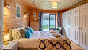 Ein gemütliches Schlafzimmer mit Holzbalkendecke, einem Bett mit Blumenmuster-Bettwäsche, einem Schminktisch und Blick ins Freie durch eine Glasdoor.