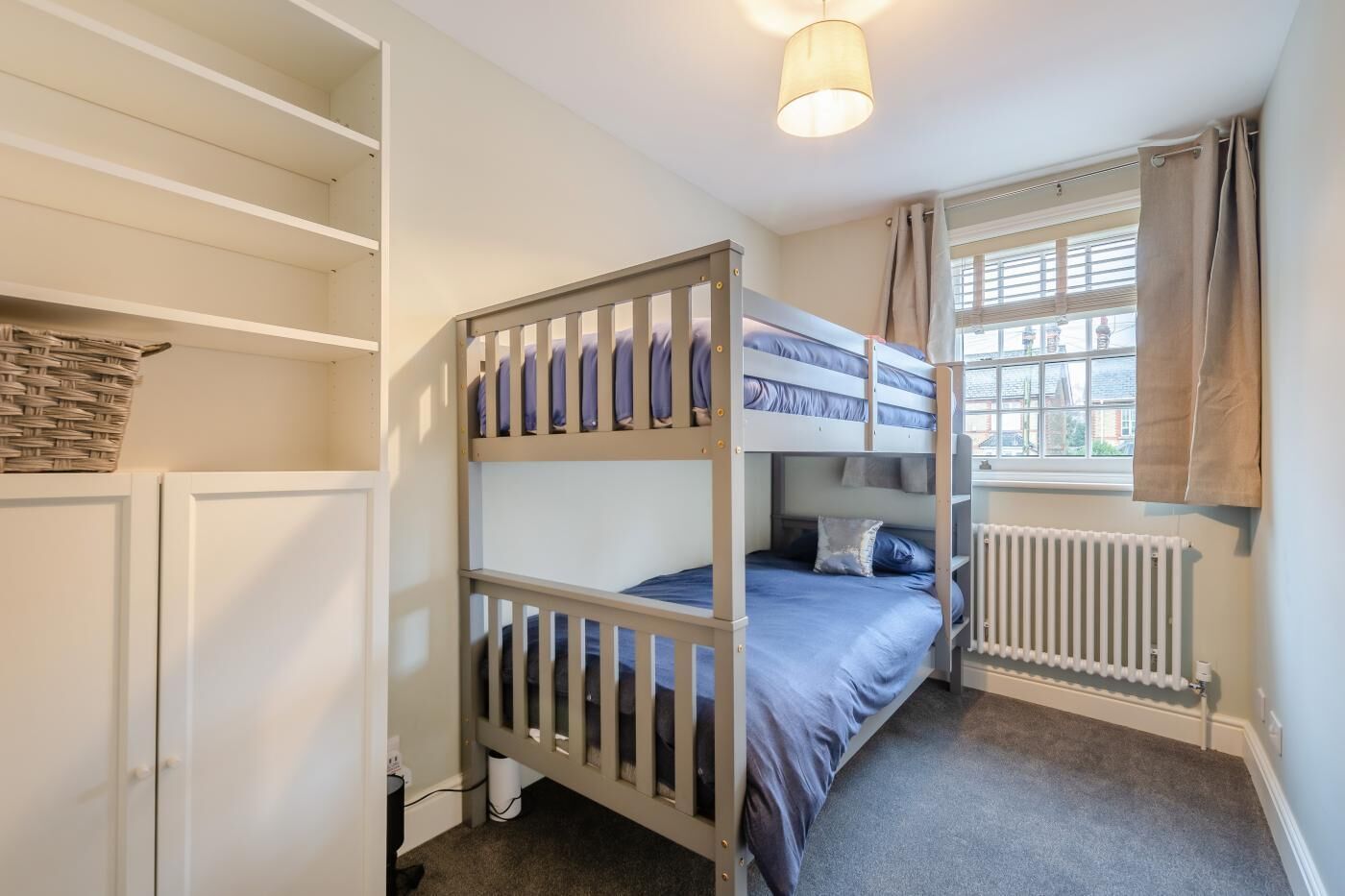 A bunk bed room with a radiator, a window with curtains, and a basket on the wall.