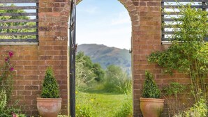 An arched brick gateway with a metal gate, surrounded by potted plants and a grassy field with mountains in the background. - 5 Bed in Church Stretton (oc-t30413) (Church Stretton)
