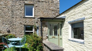 Un bâtiment en pierre avec une porte et des fenêtres vertes, un petit jardin avec des chaises et un banc en pierre.