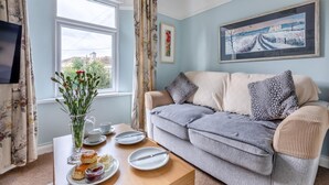 A cozy living room with a sofa, a table with a floral vase and tea set, and a painting on the wall. - 1 Bed in Teignmouth (oc-f29378) (Teignmouth)