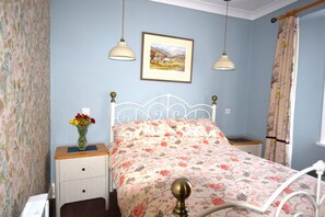 Ein Schlafzimmer mit einem Bett, zwei Nachttischen, einer Lampe, einer Vase mit Blumen, einem gerahmten Bild an der Wand und einem Vorhang.