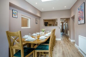 A dining area with a long wooden table set for six, flanked by wooden chairs with green cushions, a kitchen in the background, and framed pictures on the walls.