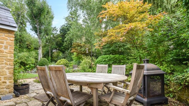 A stone patio with a round wooden table and four chairs, surrounded by lush greenery and a stone wall. - 3 Bed in Uploders (oc-g30276) (Uploders)