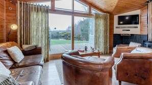 Ein gemütliches Blockhaus-Interieur mit Ledersofas, einem Fernseher und einem großen Fenster mit Blick auf eine Terrasse.