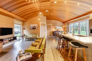 Un interior moderno con techo de madera, una sala de estar con sofá y televisor, una cocina con barra y ventanas amplias.