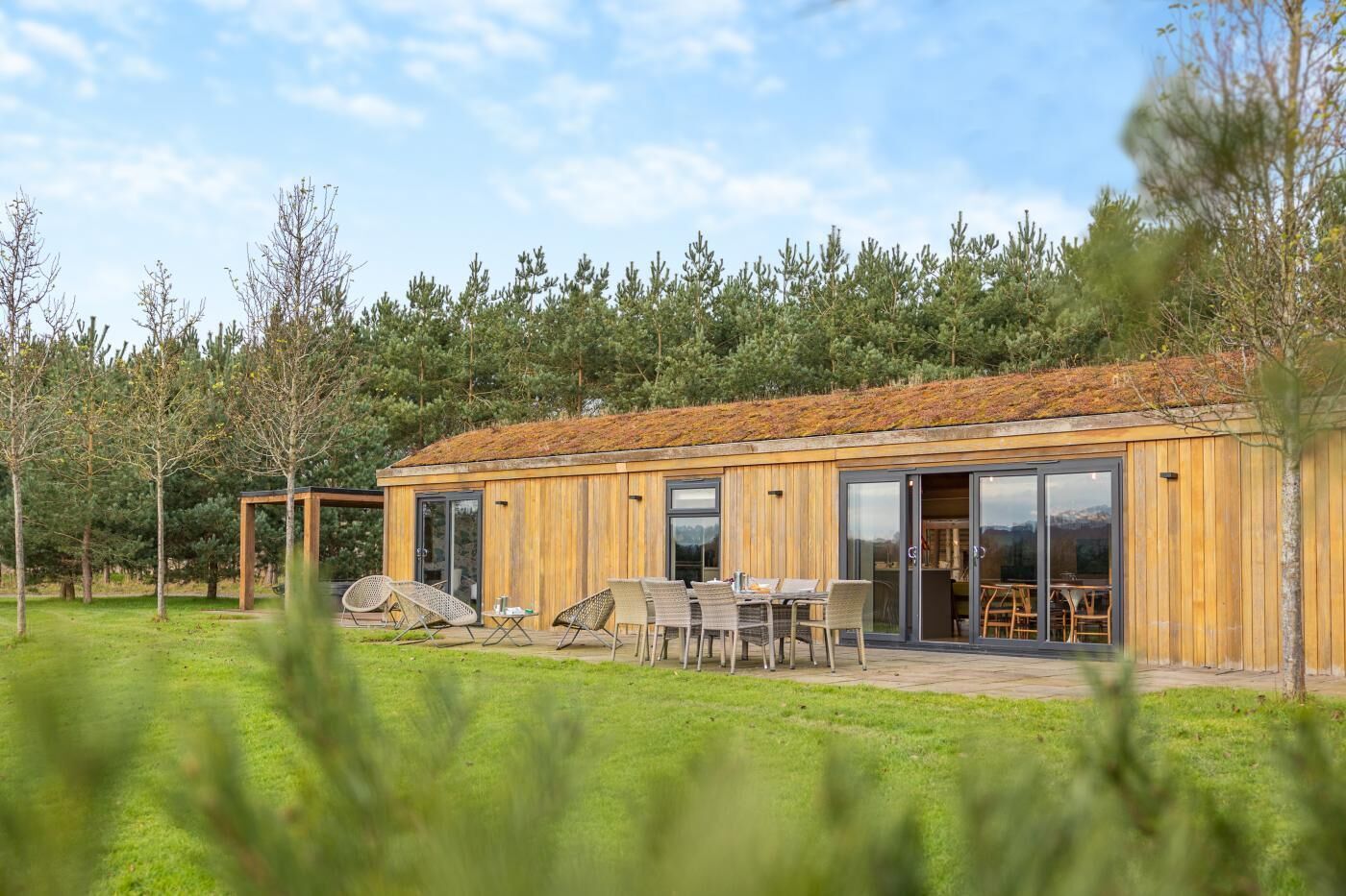 Eine Holzhütte mit Glastür, einer Terrasse mit Stühlen und Blick auf Bäume.